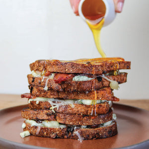 A grilled cheese sandwich made with Original Blue cheese, Toma cheese, bacon, and dried figs with honey being drizzled over the sandwich