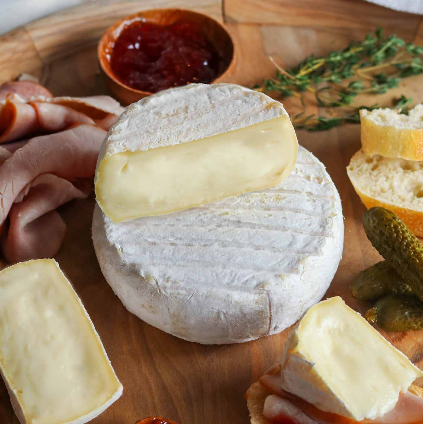 A round wheel of soft white cheese with a thick slice resting on top, arranged on a wooden board with sliced ham, bread, pickles, and a small bowl of red jam.