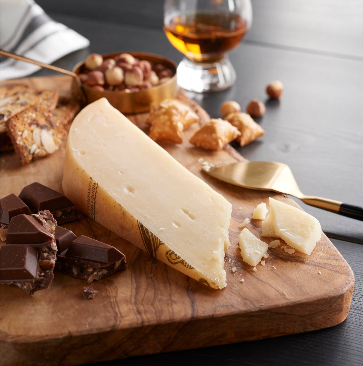 A wedge of pale yellow cheese on a wooden board with chocolate pieces, nuts, crackers, a gold cheese knife, and a glass of amber liquor on a dark tabletop.