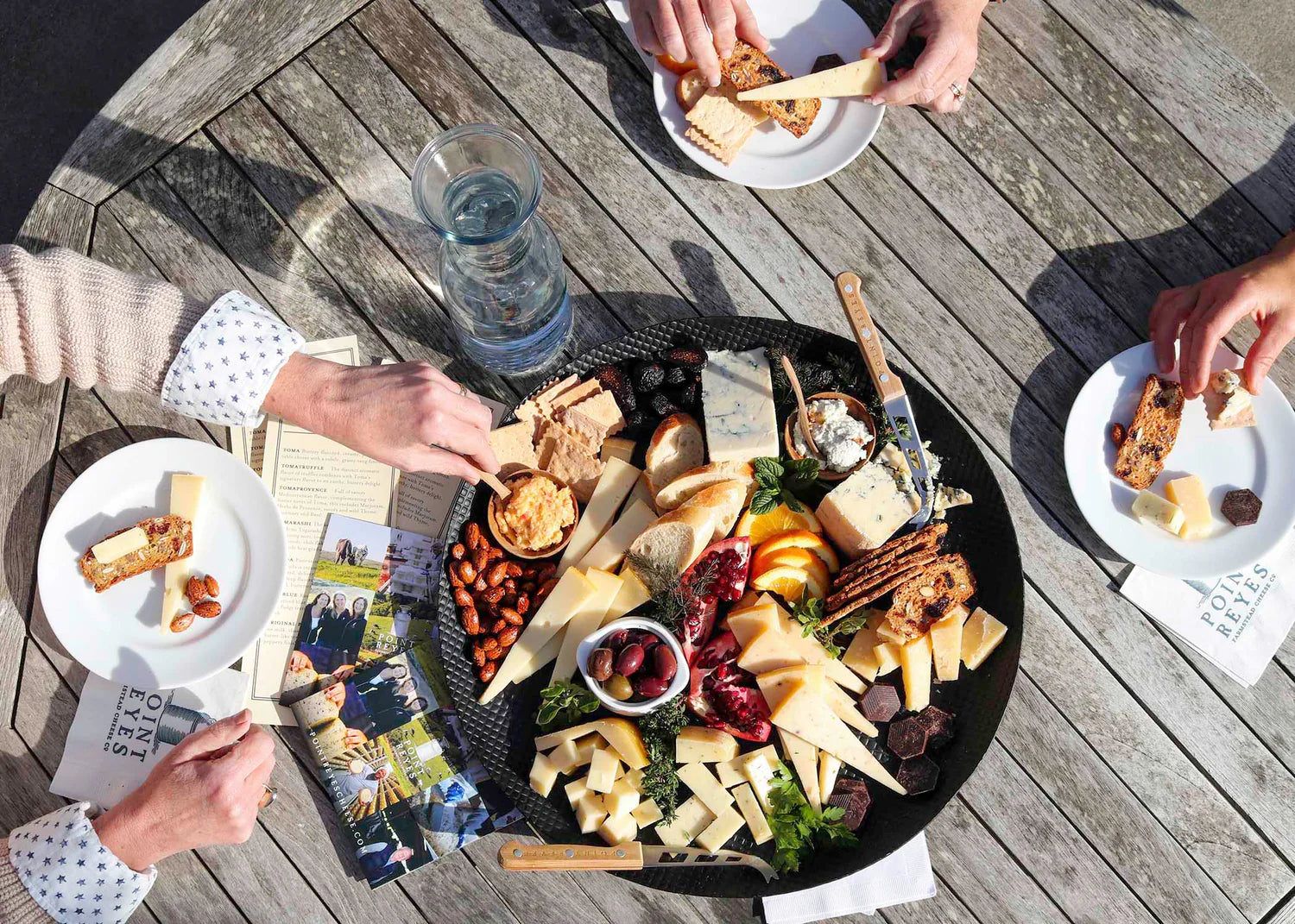 People enjoying a charcuterie board with various foods on a wooden table.